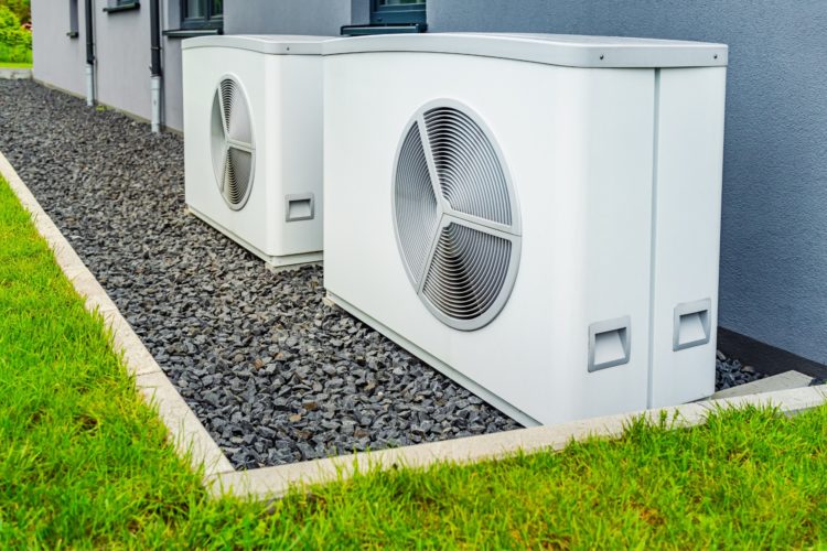 air source heat pumps, large metal boxes with fans on them, sit next to a wall on a patch of gravel. it is all surrounded by grass with a small building in the background