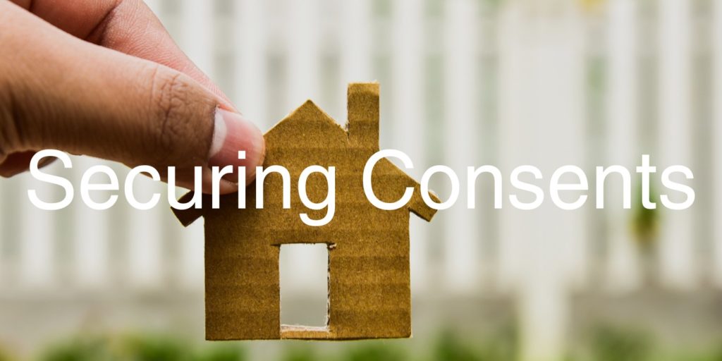 A hand holds a cardboard cutout of a house, with the word 'securing consents' in the forefront of the image